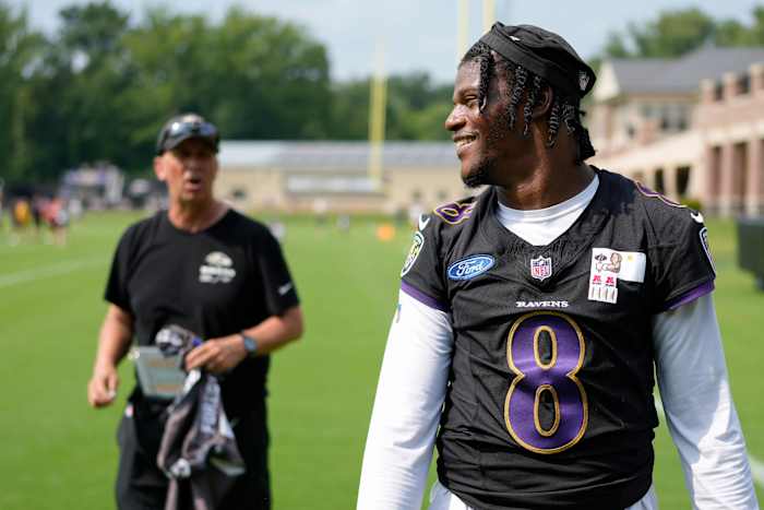 Lamar Jackson looks over his shoulder to the side and smiles with offensive coordinator Todd Monken standing in the background